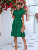 Elegant summer dress in vibrant green, stylish woman posing in front of lush floral display, straw hat and sunglasses accessorize the chic look.