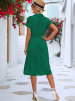 Green summer dress with waist tie detailing, worn by a woman in a scenic outdoor setting with flowering plants.