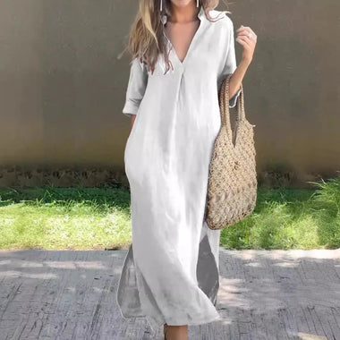 White linen maxi dress with v-neck and short sleeves, carried with a beige woven tote bag, worn outdoors on a path surrounded by greenery.