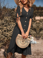 Elegant woman's black midi dress with button-down front, puff sleeves, and a stylish straw hat, holding a woven bamboo handbag in a rural field setting.