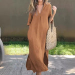 Loose cotton linen dress in warm tan color, V-neck with short sleeves, worn with woven straw tote bag on grassy outdoor setting.