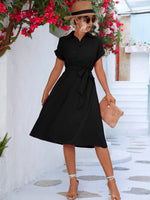 Elegant black midi dress with waist tie, woman in stylish hat and handbag walking in front of vibrant red flowers