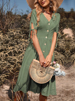 Stylish woman in green button-down midi dress, wide-brimmed sun hat, and holding a chic bamboo bag in a scenic field setting