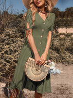 Stylish green midi dress with V-neck, button detailing, and tie sleeves for a casual summer look. Paired with a straw hat and a woven handbag, creating a chic countryside aesthetic.