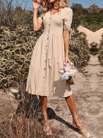 Elegant beige midi dress with button details, puff sleeves, and a flared A-line silhouette, worn by a woman in a field setting with foliage in the background.