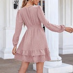 Elegant pink polka dot dress with long sleeves, V-neck, and ruffled hem, showcased against a white architectural background.